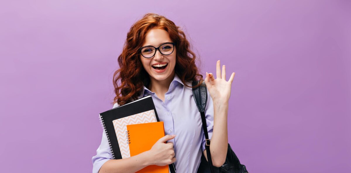 red-haired-lady-eyeglasses-holds-books-shows-ok-sign-min